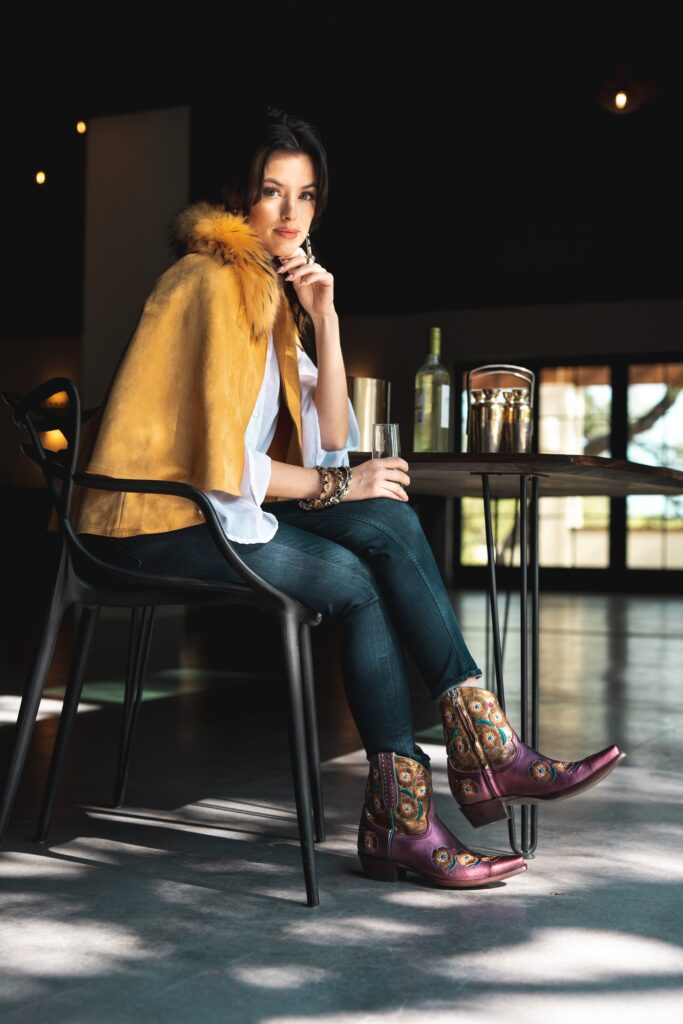 Stylish woman wearing RUJO boots and fur cape seated at modern indoor table