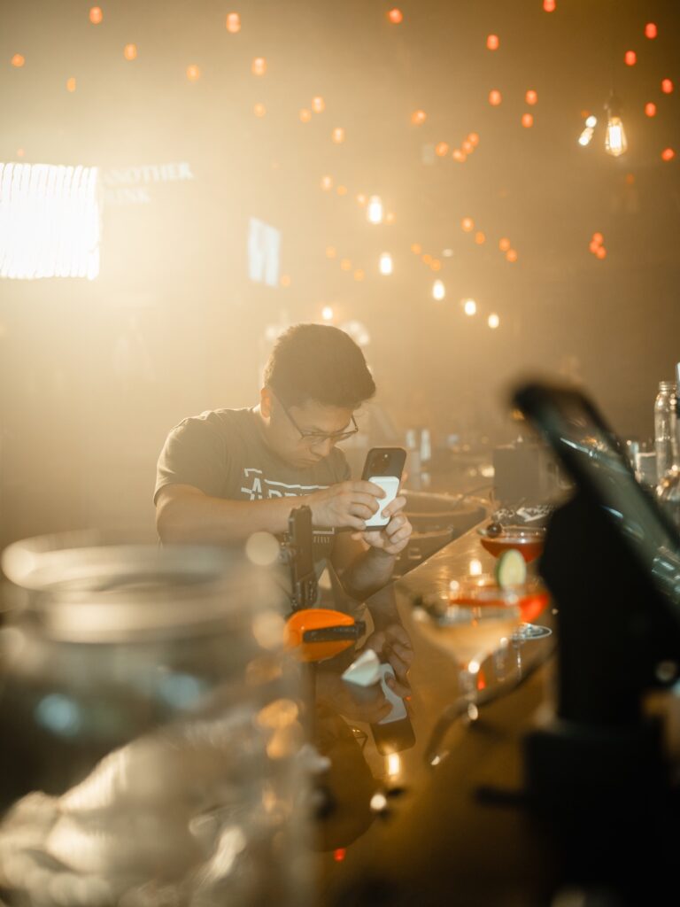 A person sits at a bar, looking at their phone, with string lights in the background.