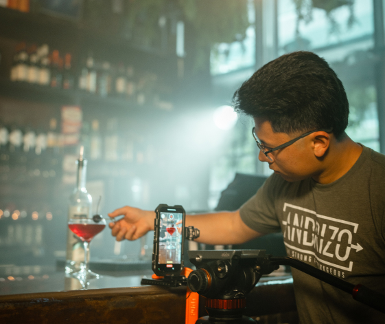 Content creator filming drink with smartphone setup at bar wearing Dazzling t-shirt