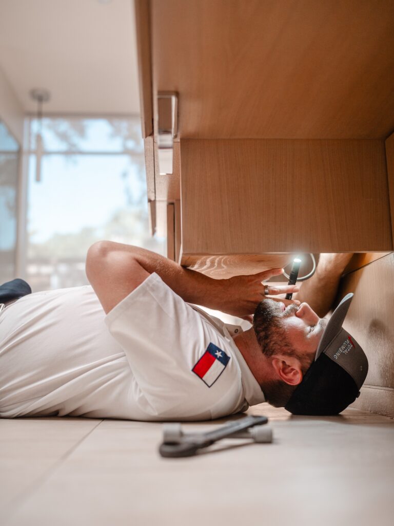 Plumber inspecting pipes under sink wearing Total Plumbing shirt and Texas flag patch