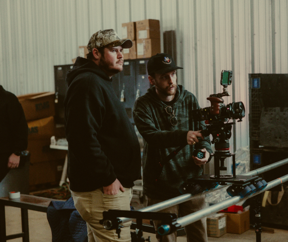A man in a plaid shirt operates a professional video camera on a dolly while another man watches.