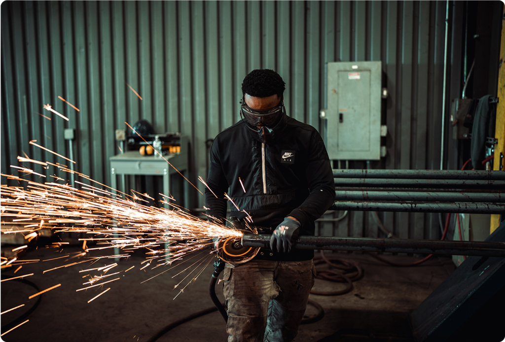 A worker in a workshop uses an angle grinder on a metal pipe, creating a shower of sparks.