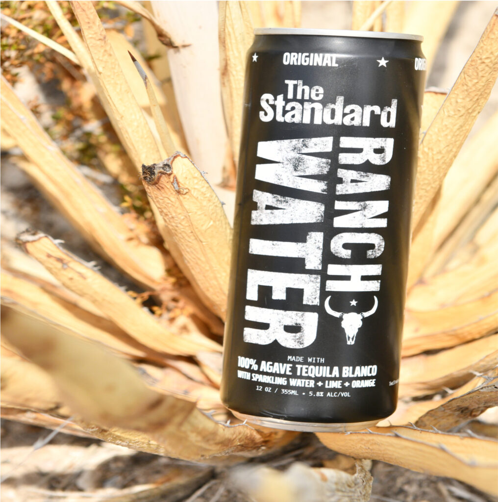 A black can of The Standard Ranch Water is shown among the leaves of a dry plant.